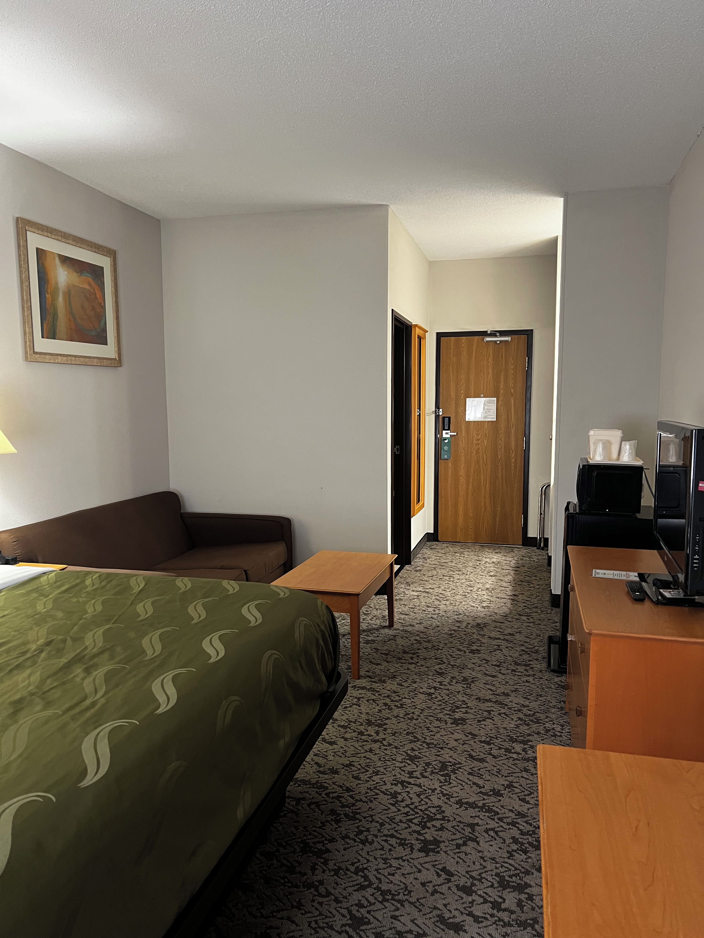 A hotel room with a bed, a desk, a chair, a sofa, a television, and a door leading to another room.