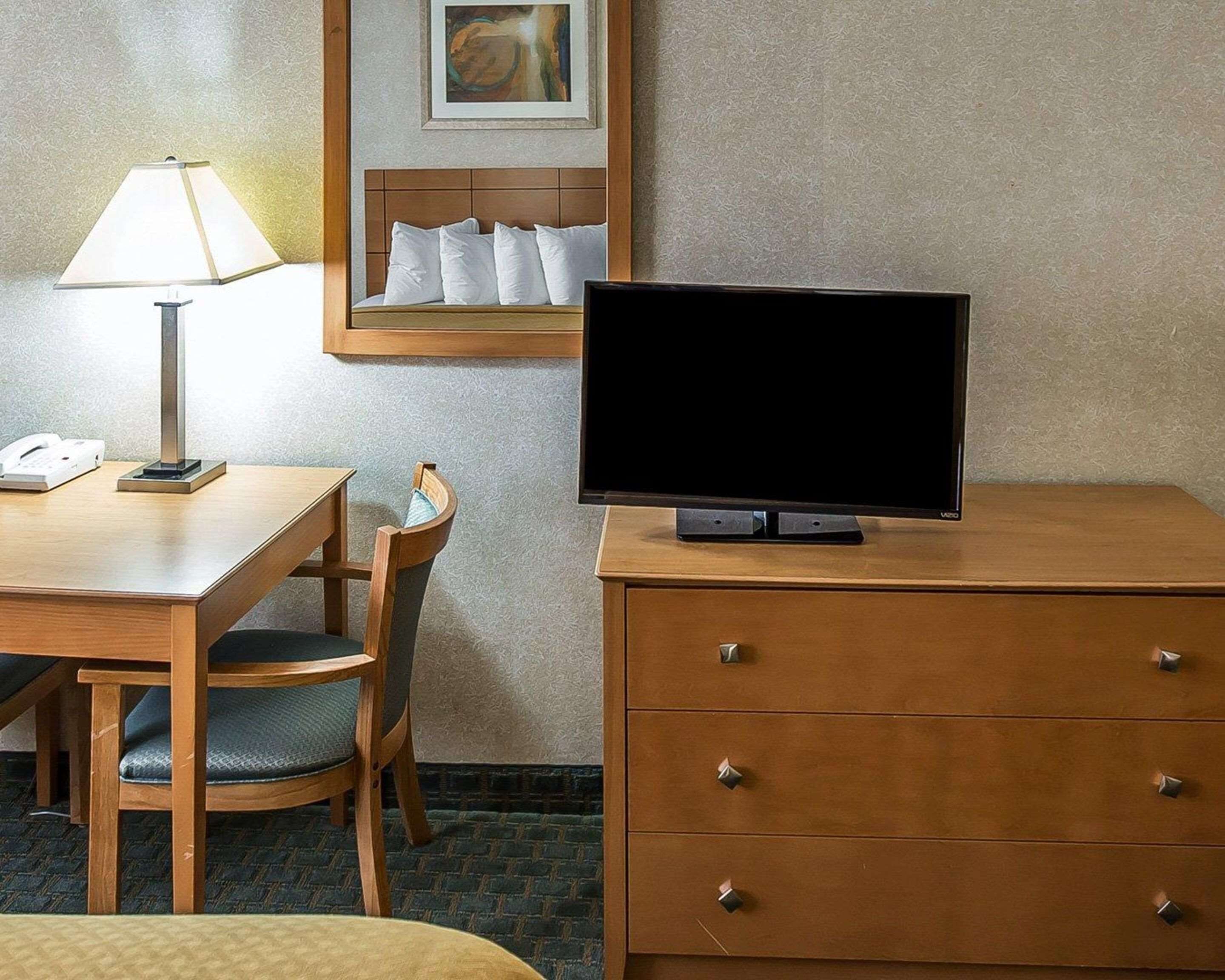 Une chambre d’hôtel avec un bureau en bois, une chaise, un téléviseur posé sur une commode en bois, une lampe et une œuvre d’art encadrée accrochée au mur.