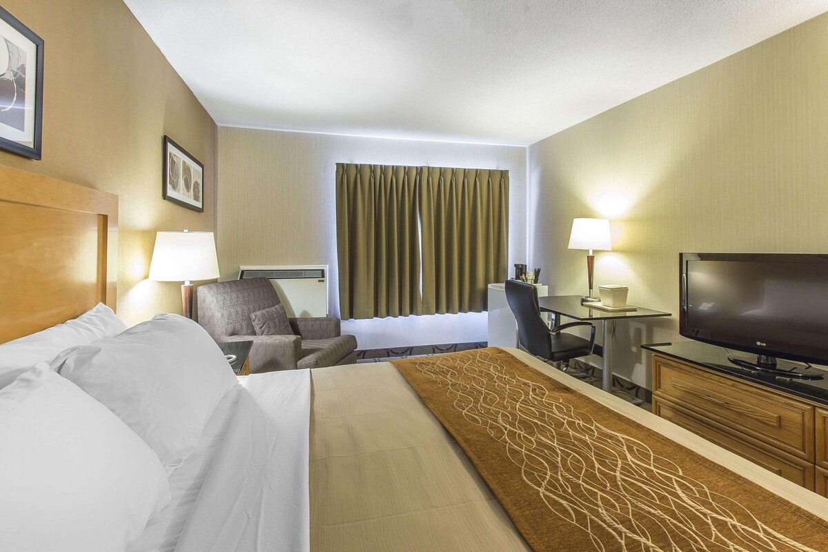 A hotel room with a large bed, a desk with a television, a chair, and a lamp.