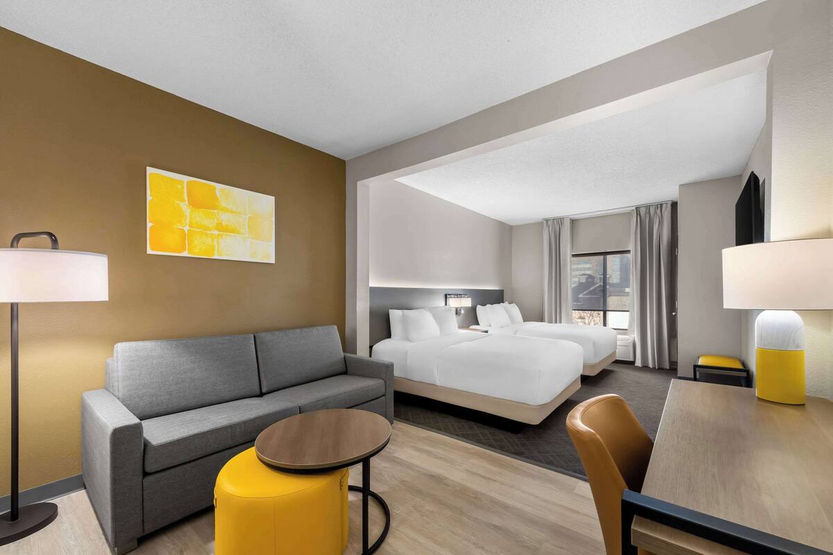 A hotel room with a grey sofa, a bed, a small round table, and a yellow ottoman.