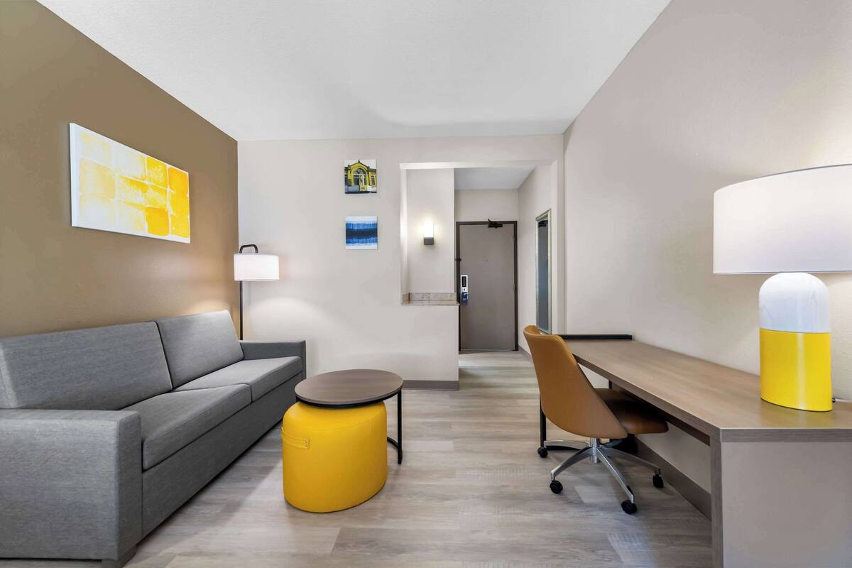 A modern hotel room with a grey sofa, a yellow ottoman, a wooden desk, and a chair.