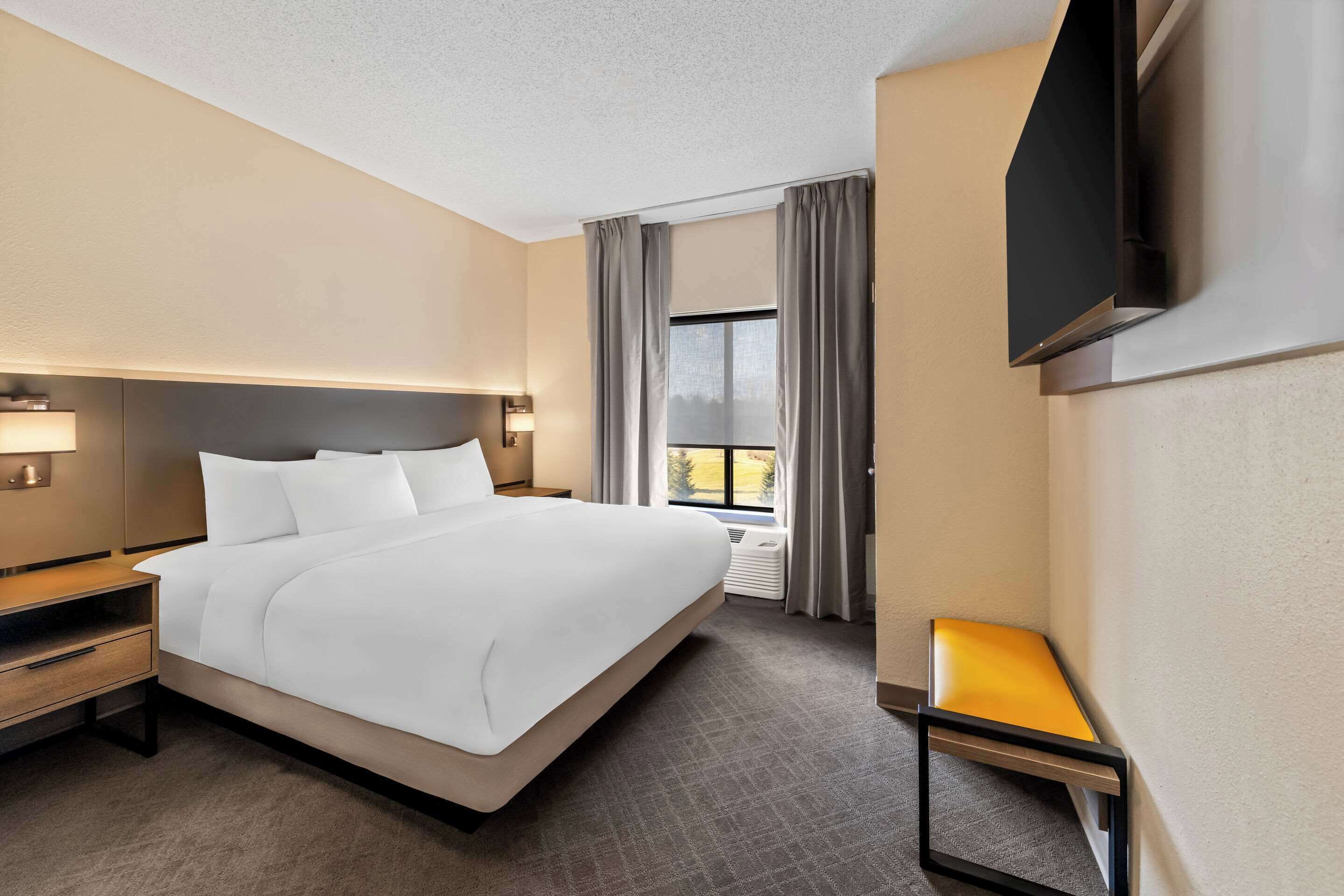 A hotel room with a large bed, a bedside table with a lamp, a small table with a chair, a TV mounted on the wall, and a window with curtains.
