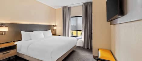 A hotel room with a large bed, a bedside table with a lamp, a small table with a chair, a TV mounted on the wall, and a window with curtains.