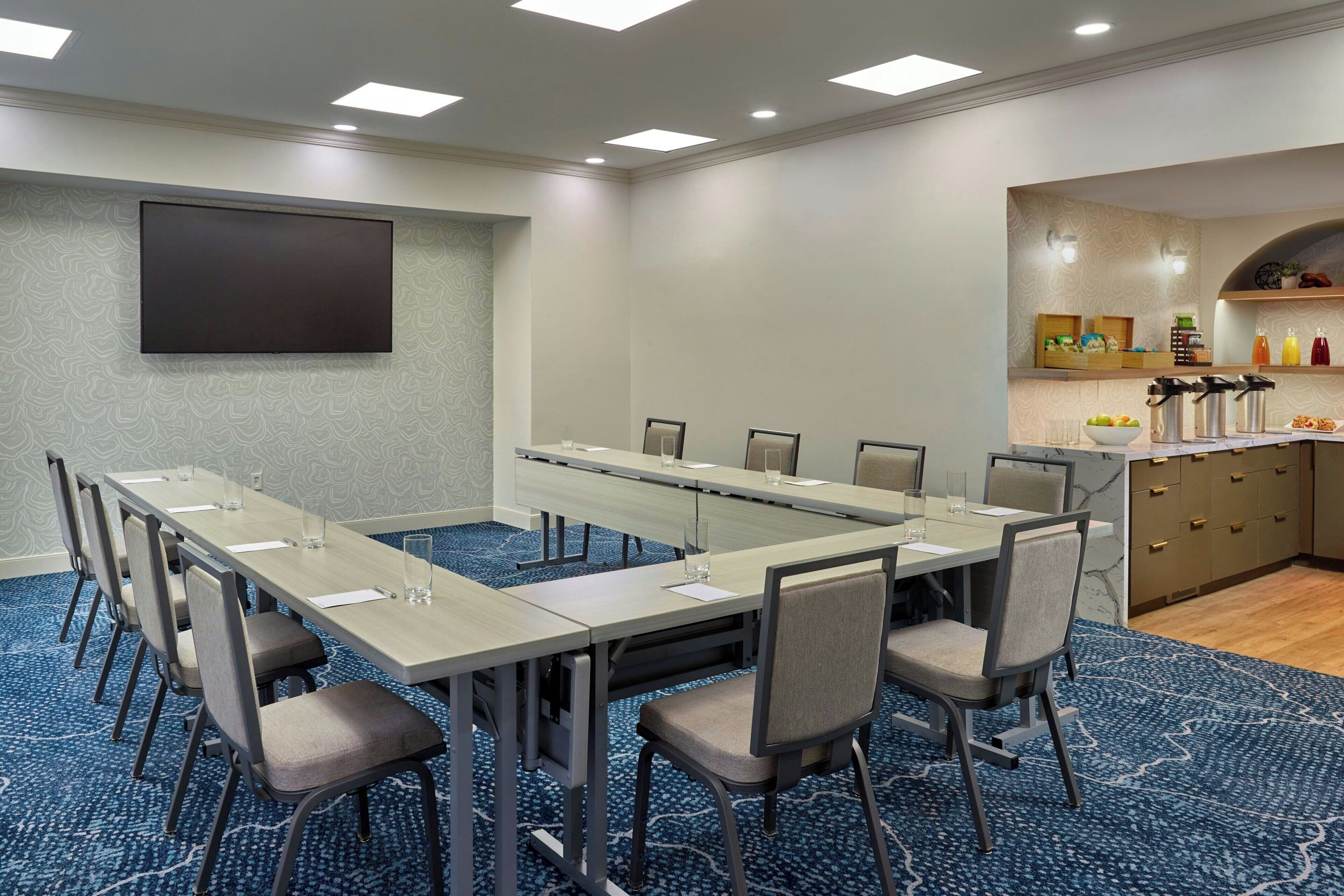 Un salón de conferencias con una larga mesa, sillas, un televisor de pantalla plana y una zona de cocina con estantes y electrodomésticos.