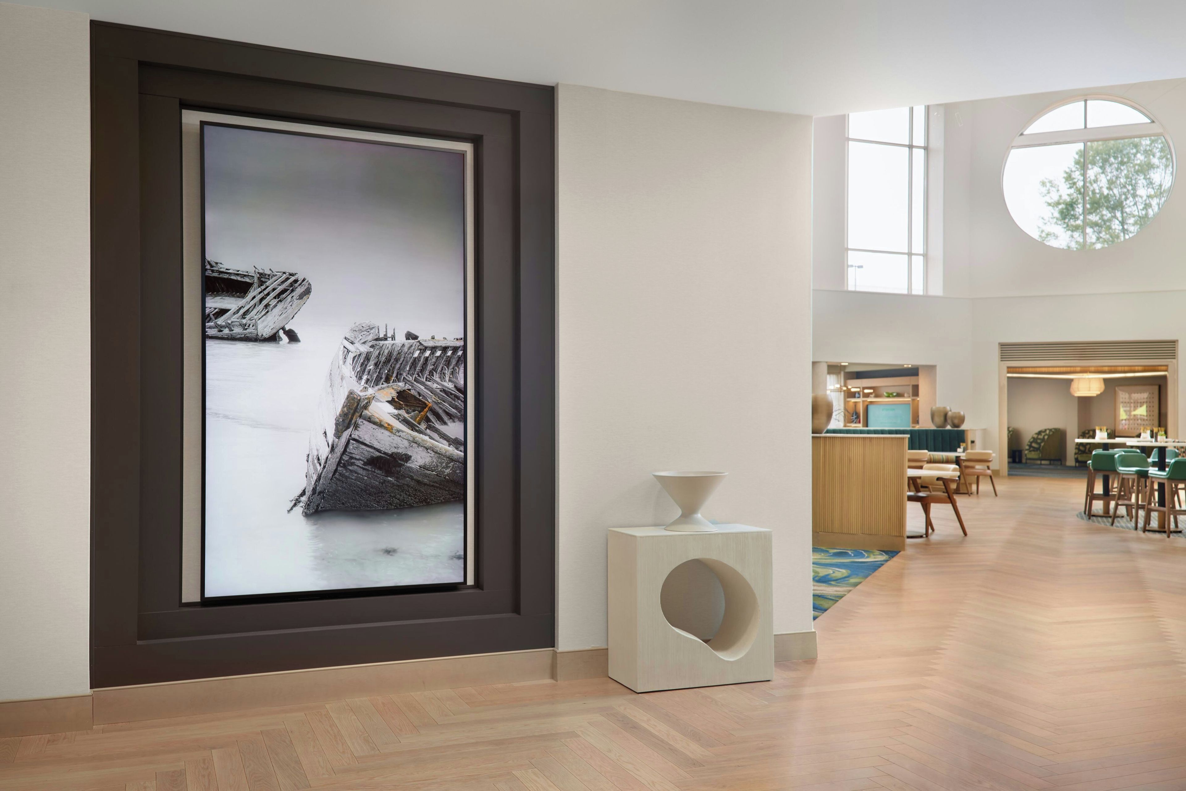 A modern hotel lobby with a large framed artwork of a shipwreck, a reception desk, and a dining area with chairs and tables.