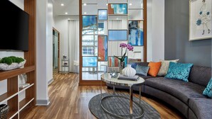 A modern living room with a grey sofa, a round coffee table, and a TV on the left. There is a large window with curtains, a bookshelf, and a decorative wall panel. - Renaissance Charlotte SouthPark (Charlotte)