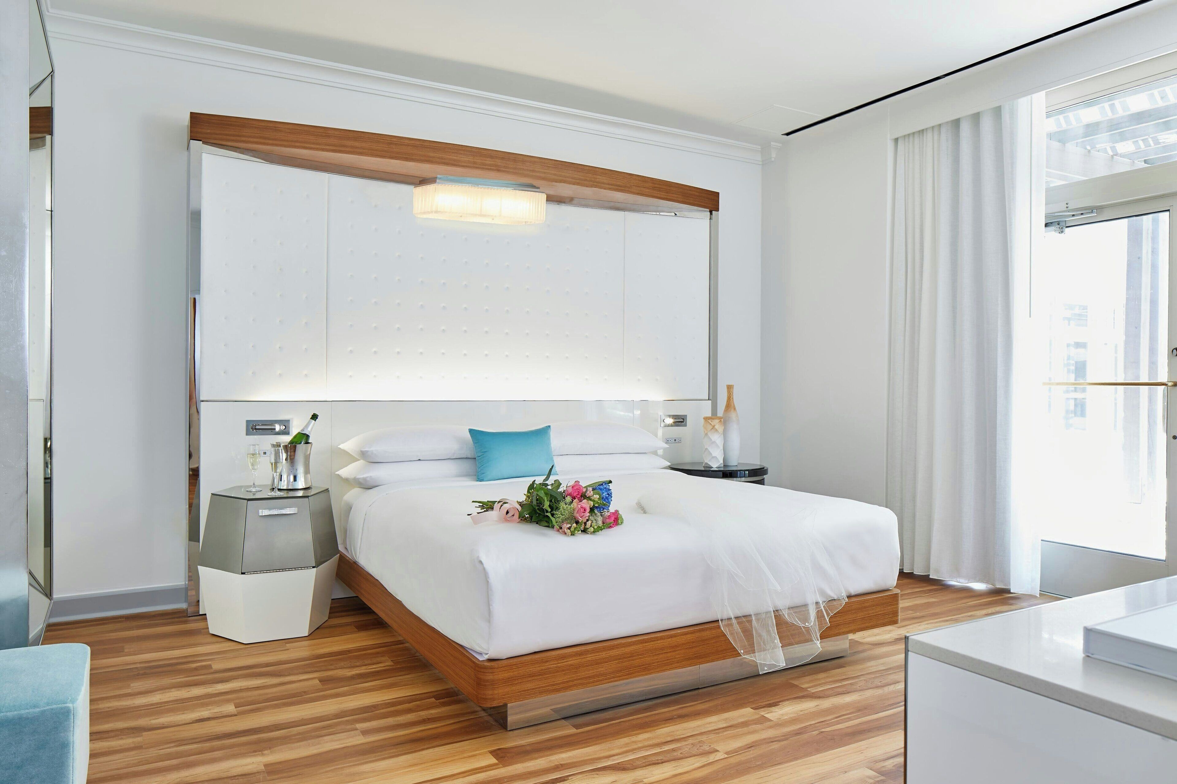 A modern hotel room with a large bed, wooden headboard, bedside tables, and a large window with curtains.