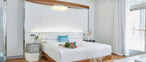 A modern hotel room with a large bed, wooden headboard, bedside tables, and a large window with curtains.