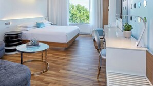 Habitación de hotel moderna con cama, escritorio, silla, mesa redonda y un ventanal con vistas a árboles.