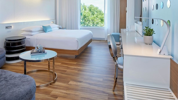 Ein modernes Hotelzimmer mit Bett, Schreibtisch, Stuhl, rundem Tisch und einem großen Fenster mit Blick auf Bäume.