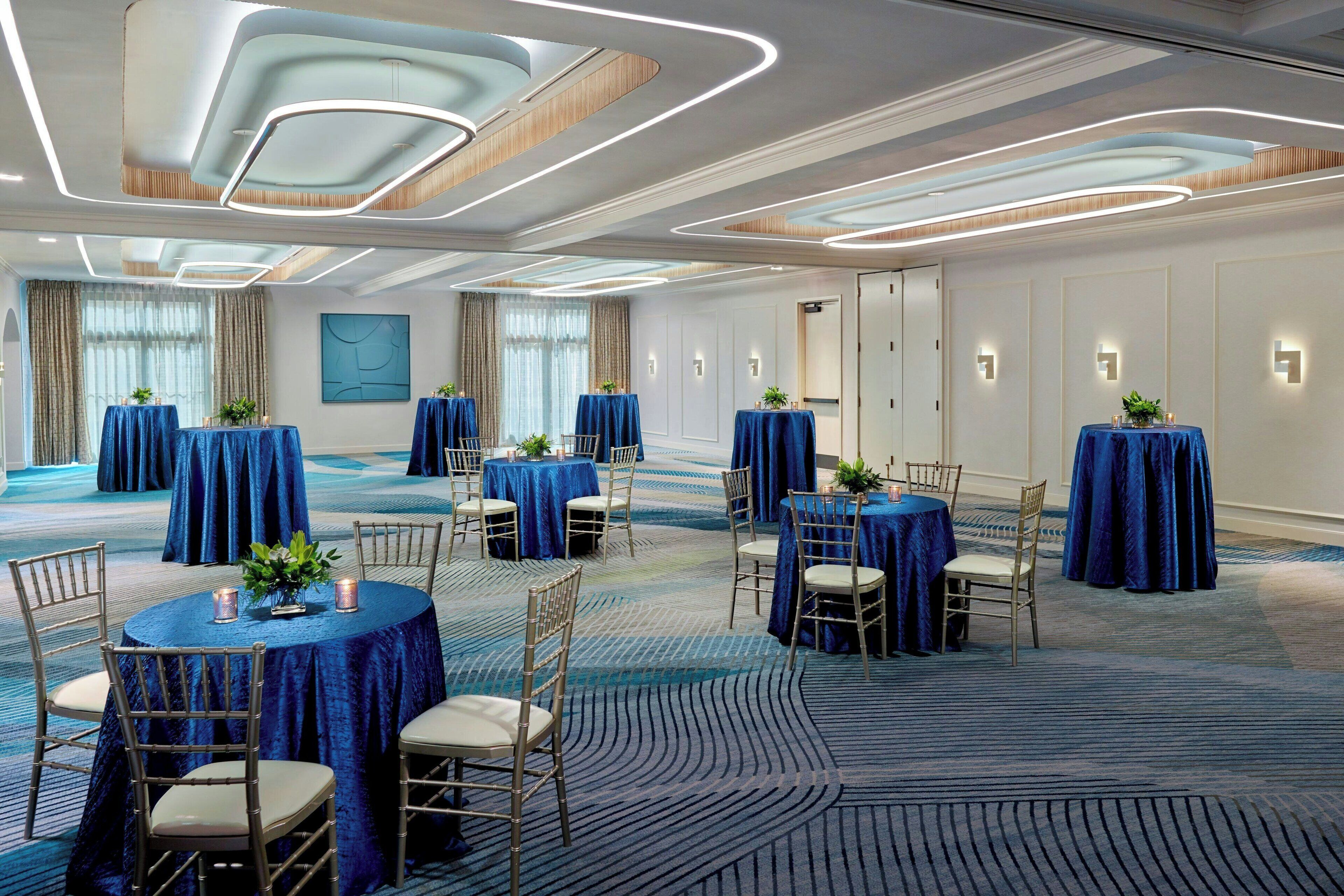 A hotel conference room with round tables covered in blue tablecloths, gold chairs, and decorative plants.