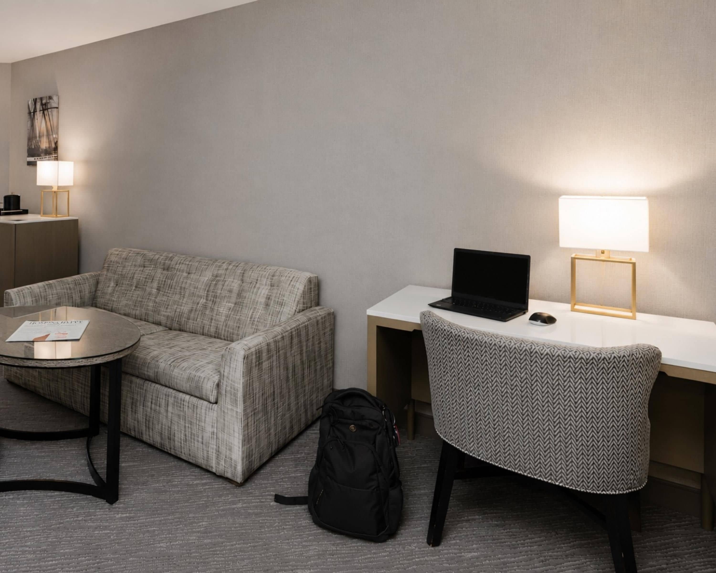A hotel room with a sofa, a small round table, a desk with a laptop, a chair, a lamp, and a backpack on the floor.