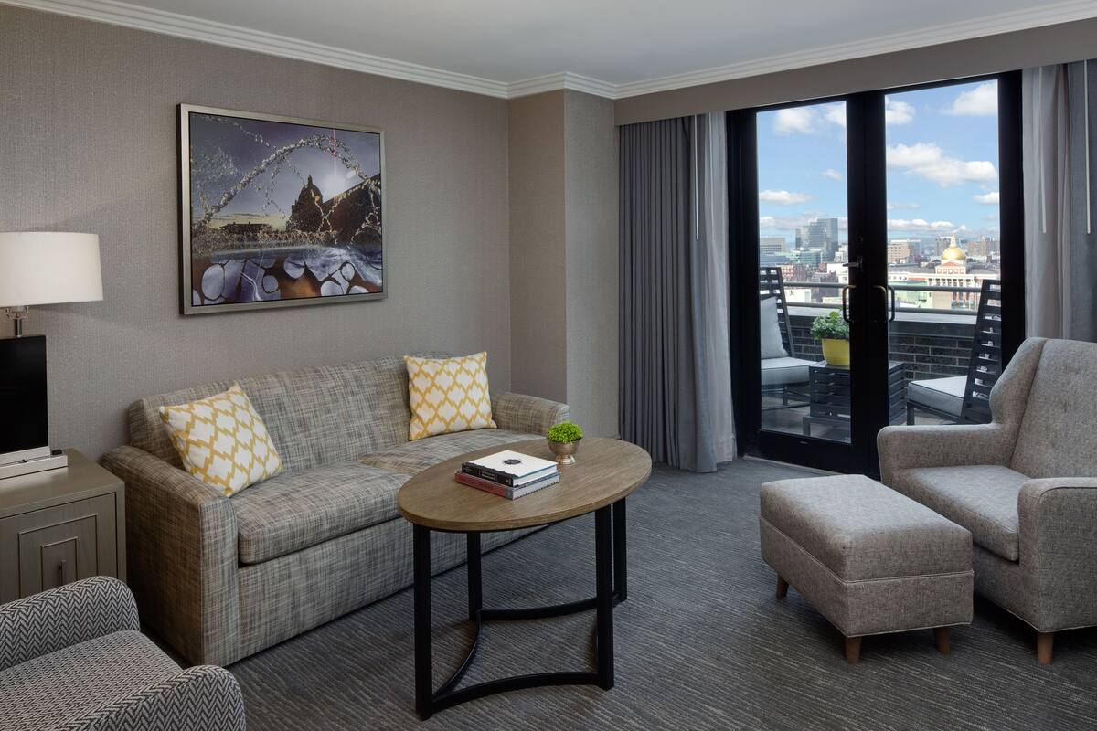 A modern hotel room with a grey sofa, a matching armchair, a small round table with a book and a plant, and a large window with a city view.