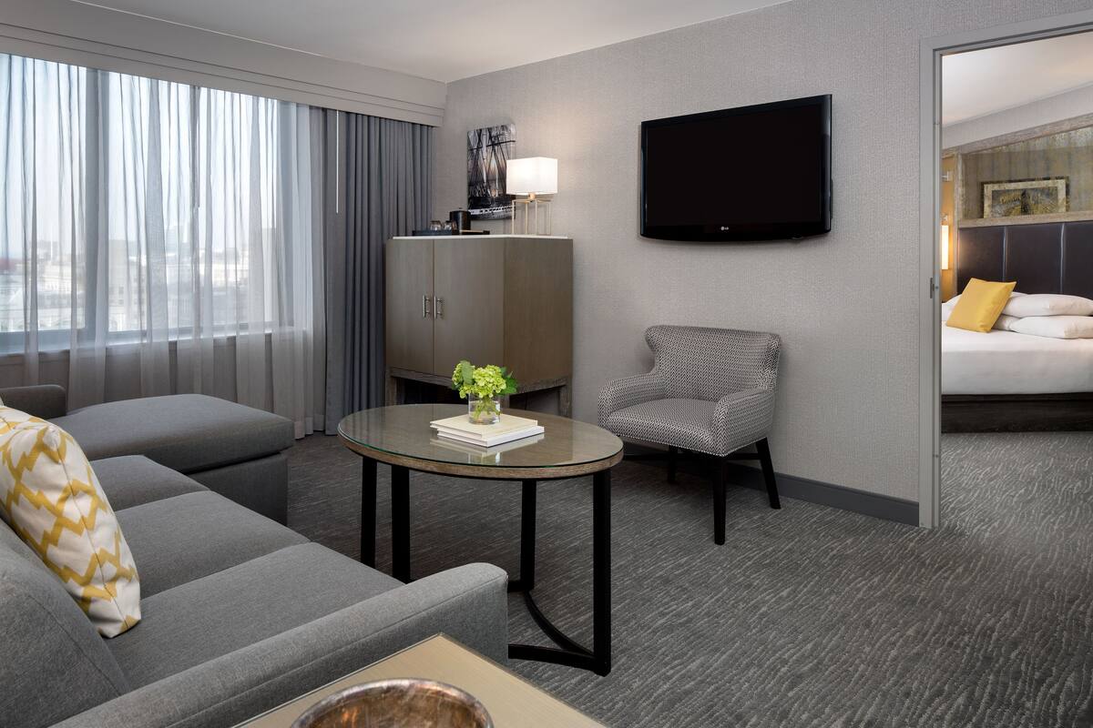 A modern hotel room with a grey sofa, a small round table with a vase, a grey armchair, a bed with a yellow pillow, a TV mounted on the wall, and a large window with curtains.