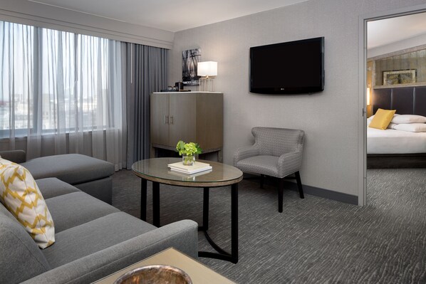 A modern hotel room with a grey sofa, a small round table with a vase, a grey armchair, a bed with a yellow pillow, a TV mounted on the wall, and a large window with curtains.