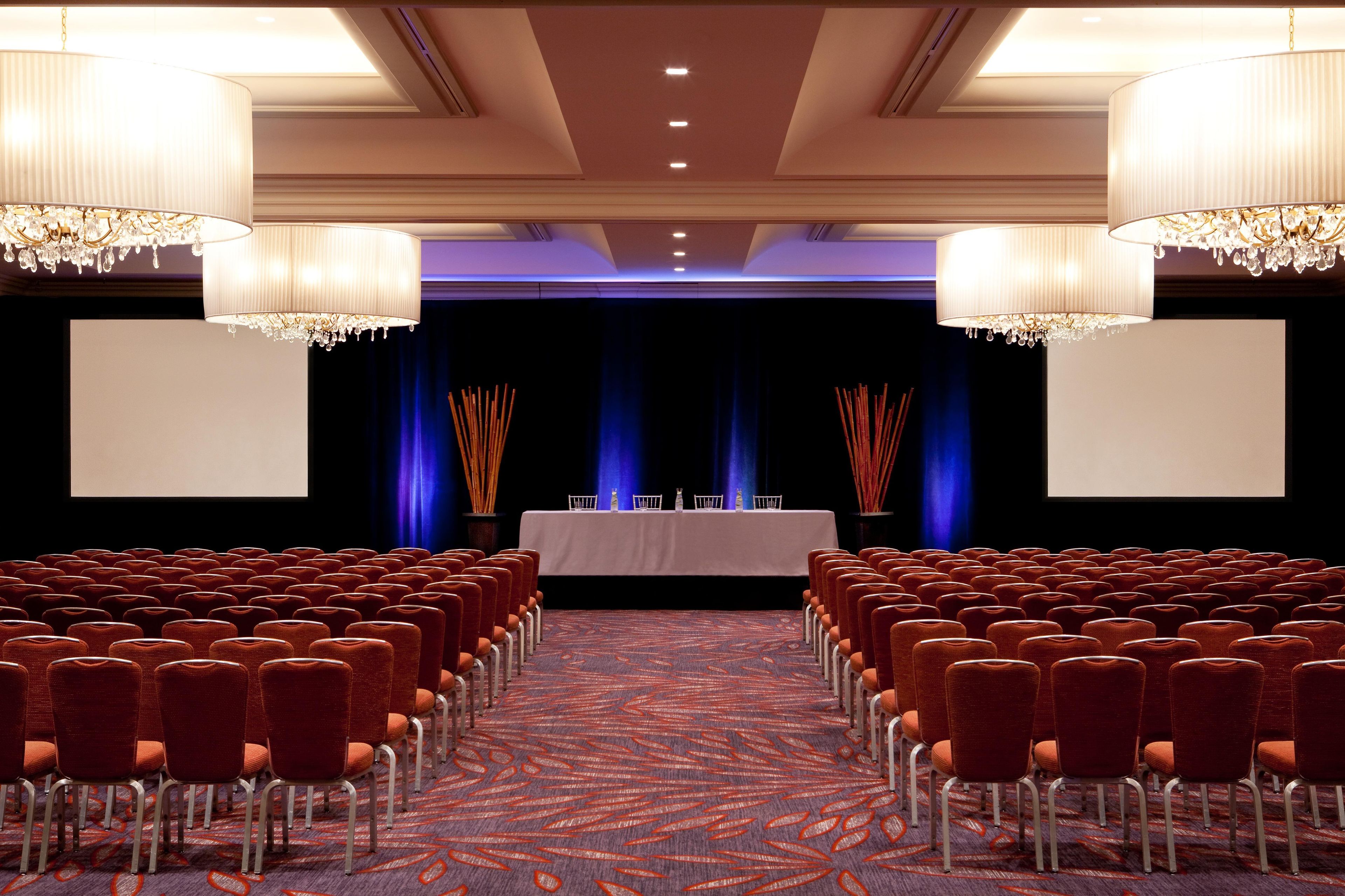 A conference room with rows of chairs, a stage with a table and chairs, and chandeliers.