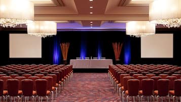 Un salón de conferencias con filas de sillas, un escenario con una mesa y sillas, y candelabros.