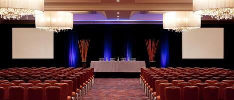 A conference room with rows of chairs, a stage with a table and chairs, and chandeliers.