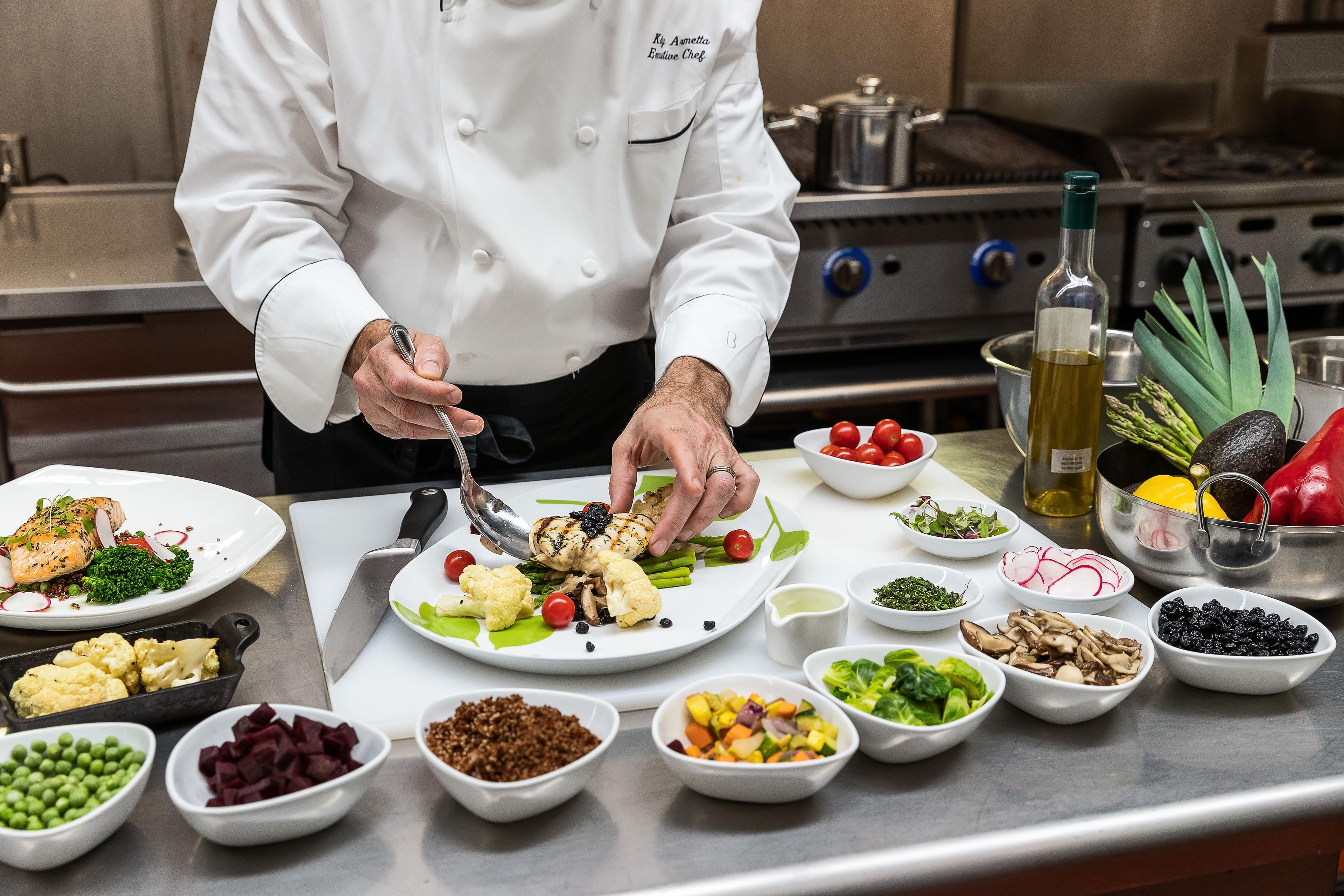 Un chef preparando un plato con diversos vegetales y mariscos sobre una encimera de acero inoxidable en una cocina.