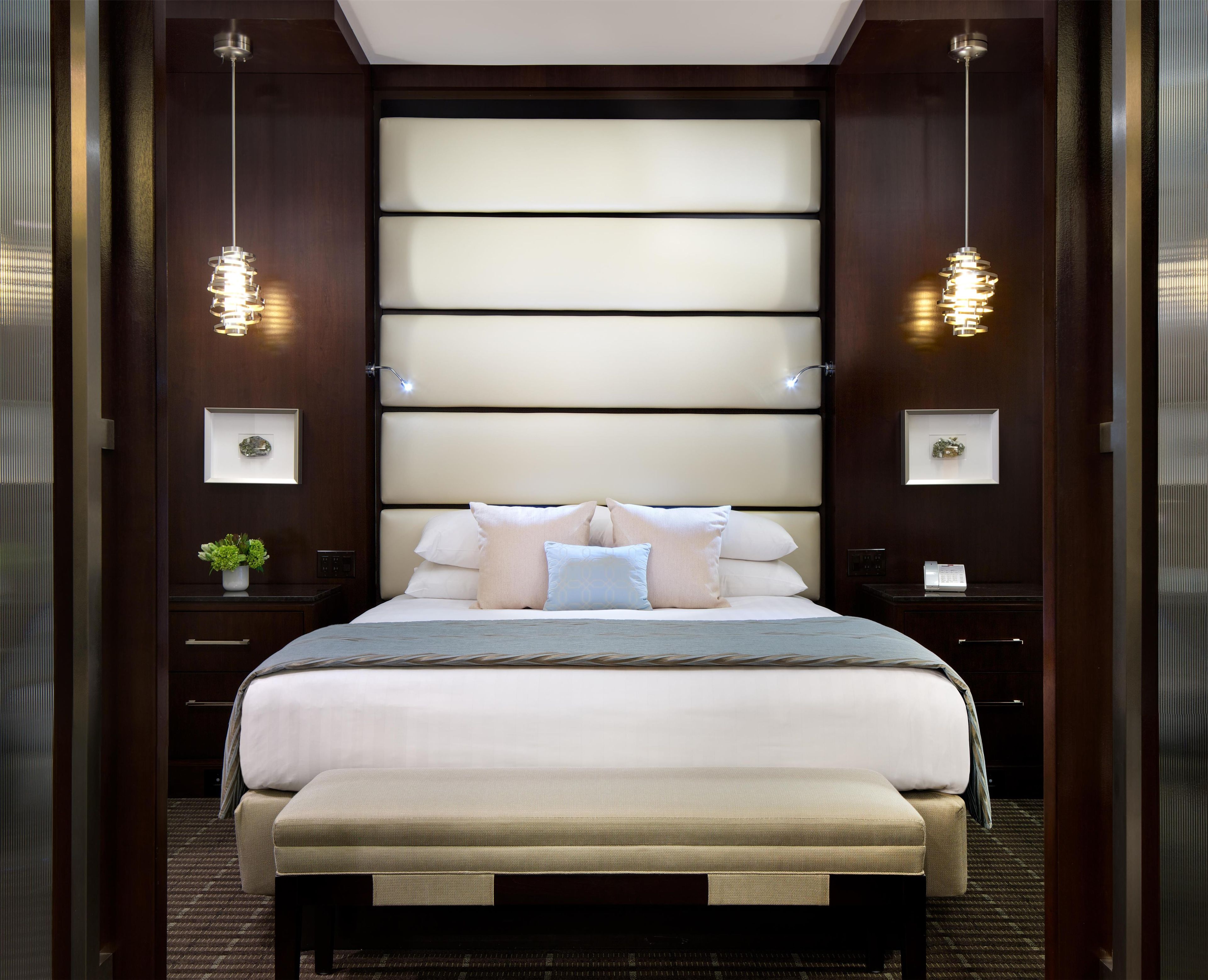 Una habitación de hotel con una cama grande, mesitas de noche, un banco e iluminación en la pared.