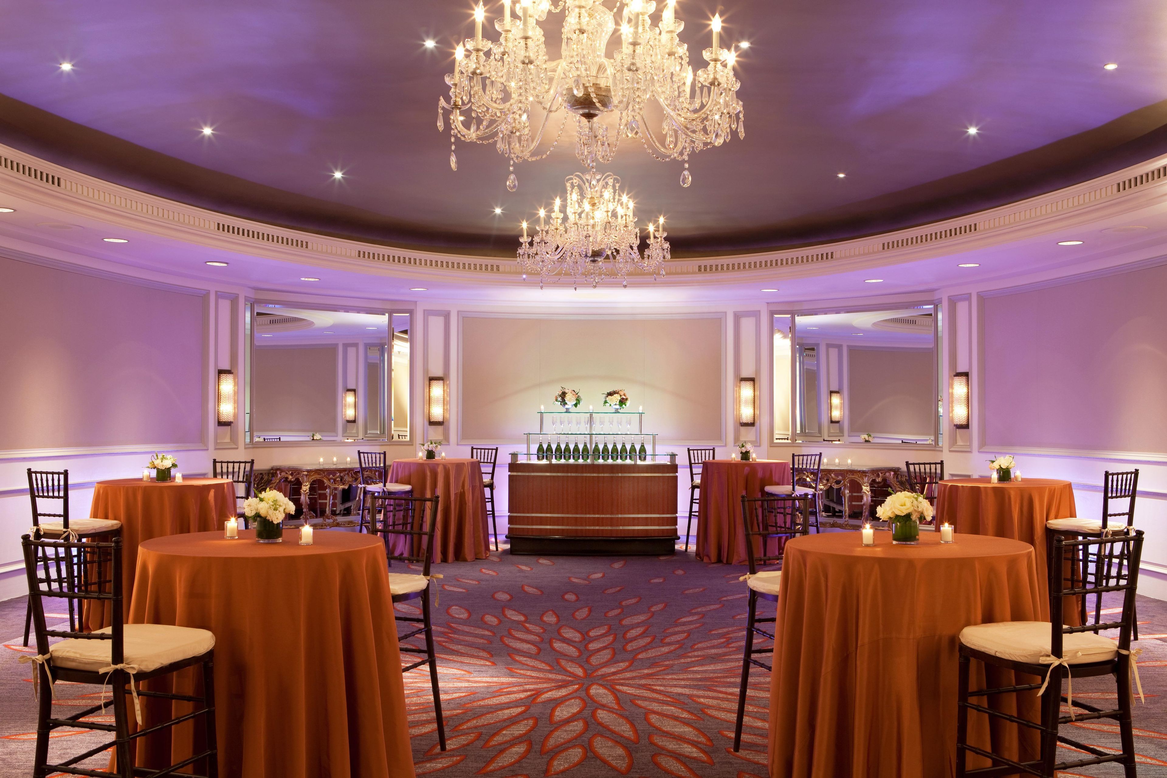 A hotel conference room with round tables, chairs, a bar area, and a large chandelier.