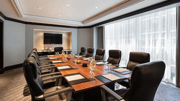 Un salón de conferencias con una larga mesa, varias sillas, un televisor de pantalla plana y ventanas amplias.