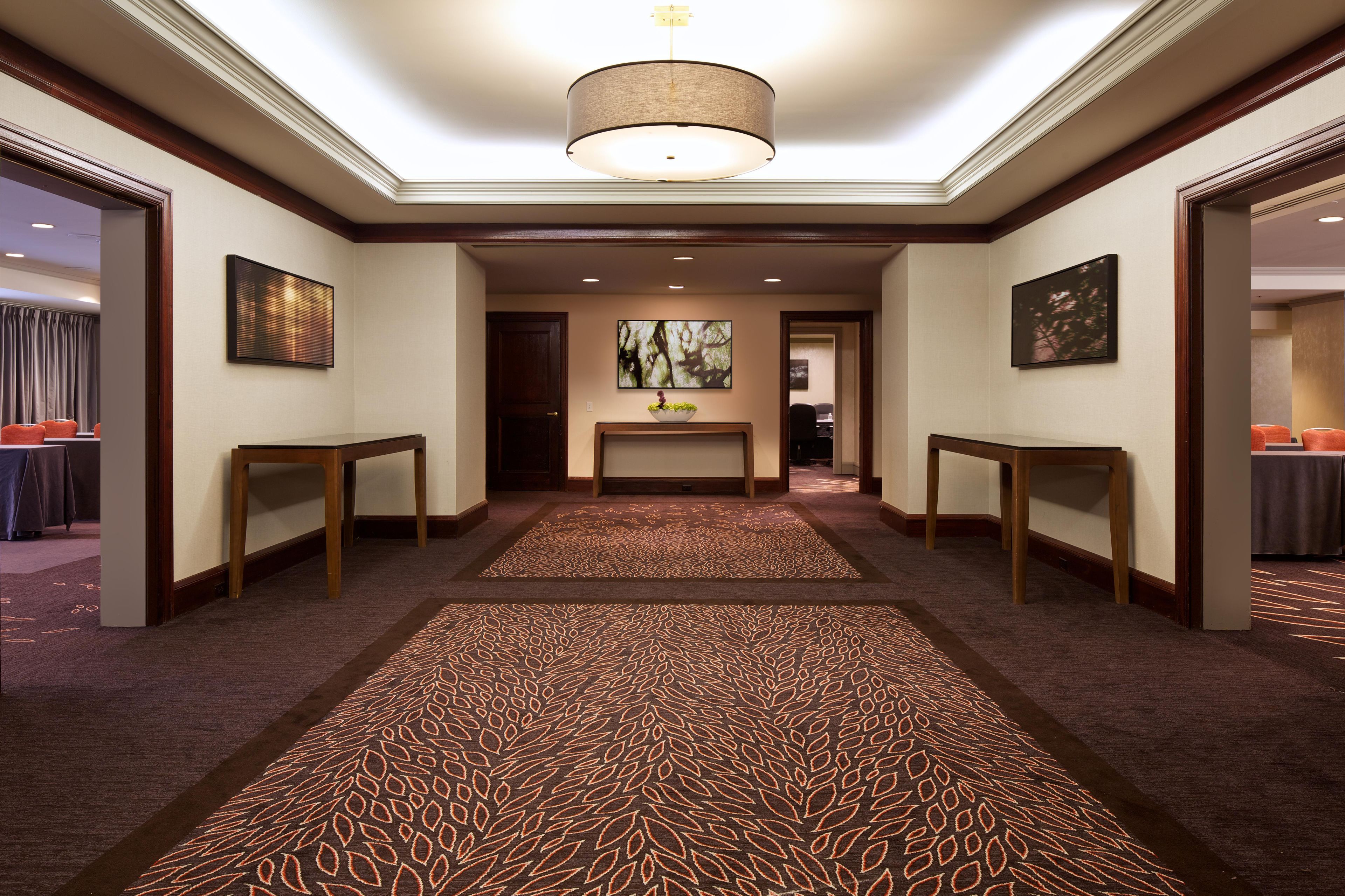 Un pasillo de hotel con alfombra estampada, mesas de madera y obras de arte en las paredes.