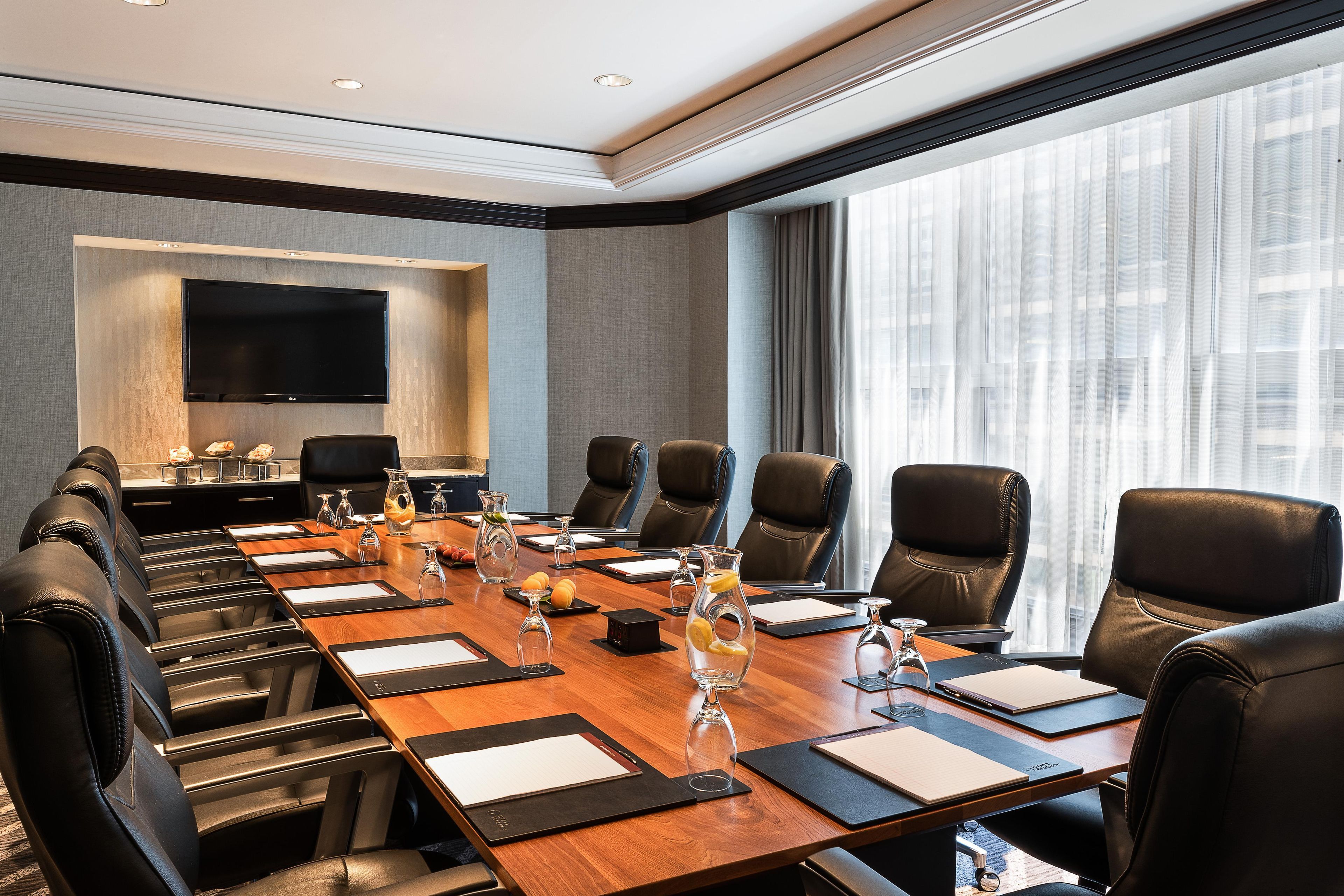 Une salle de conférence avec une longue table, de nombreuses chaises, un téléviseur à écran plat et une grande fenêtre avec des rideaux.