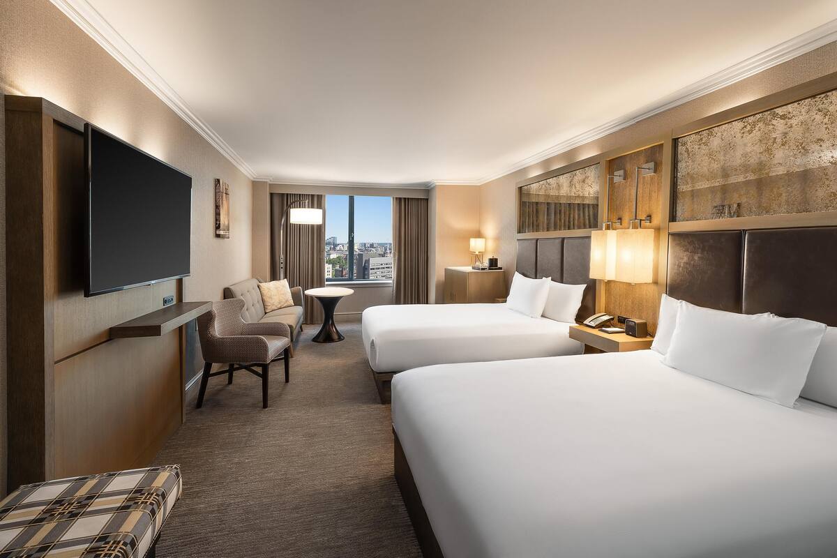 A hotel room with two beds, a flat-screen TV, a desk, a chair, and a view of the city.