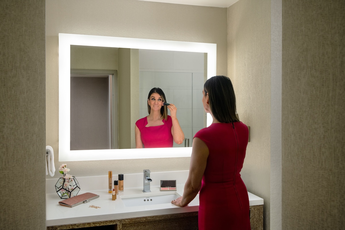 A woman in a red dress standing in front of a bathroom mirror with a white countertop, a vanity, and a door in the background.