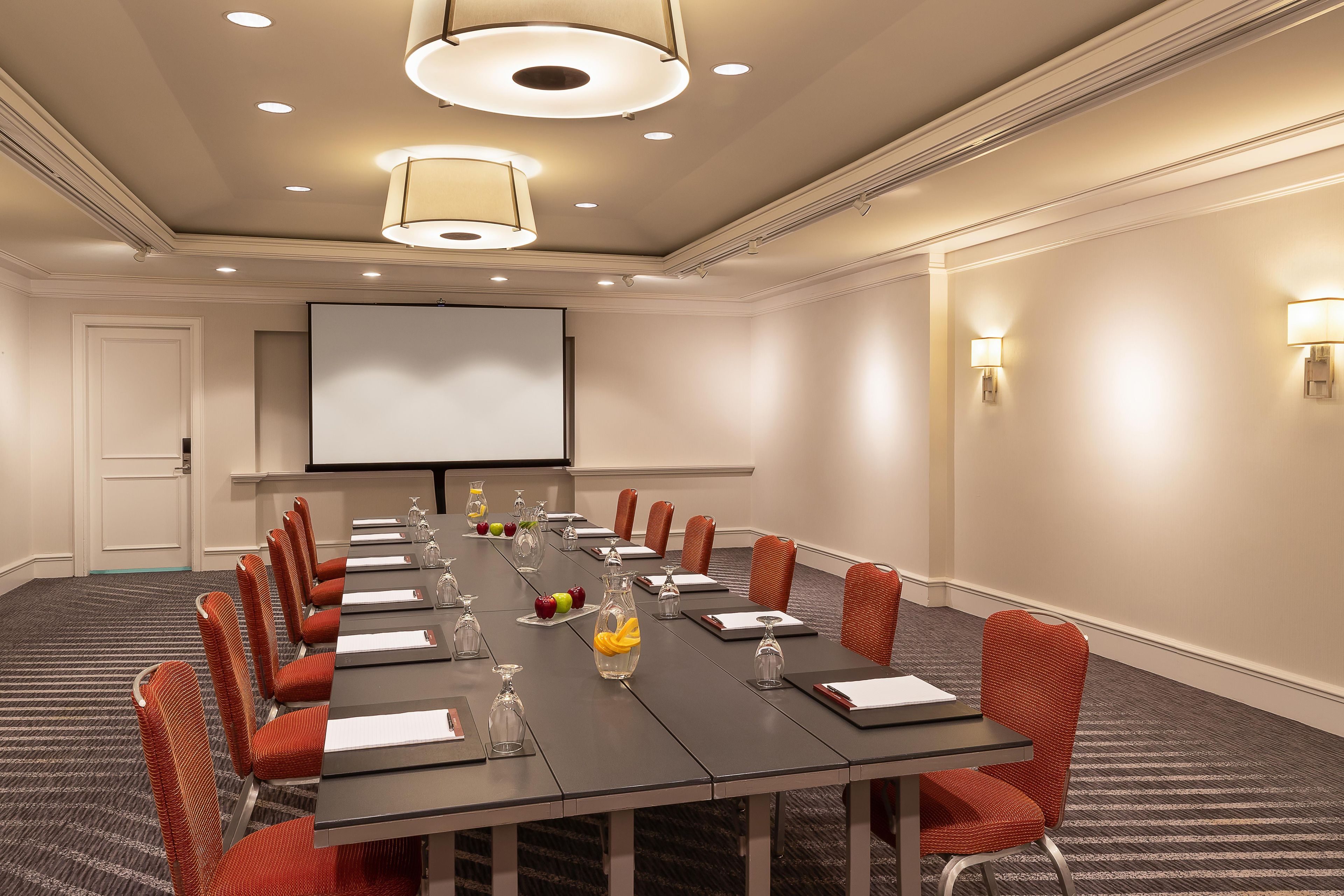 A conference room with a long table, chairs, a projector screen, and a carpeted floor.