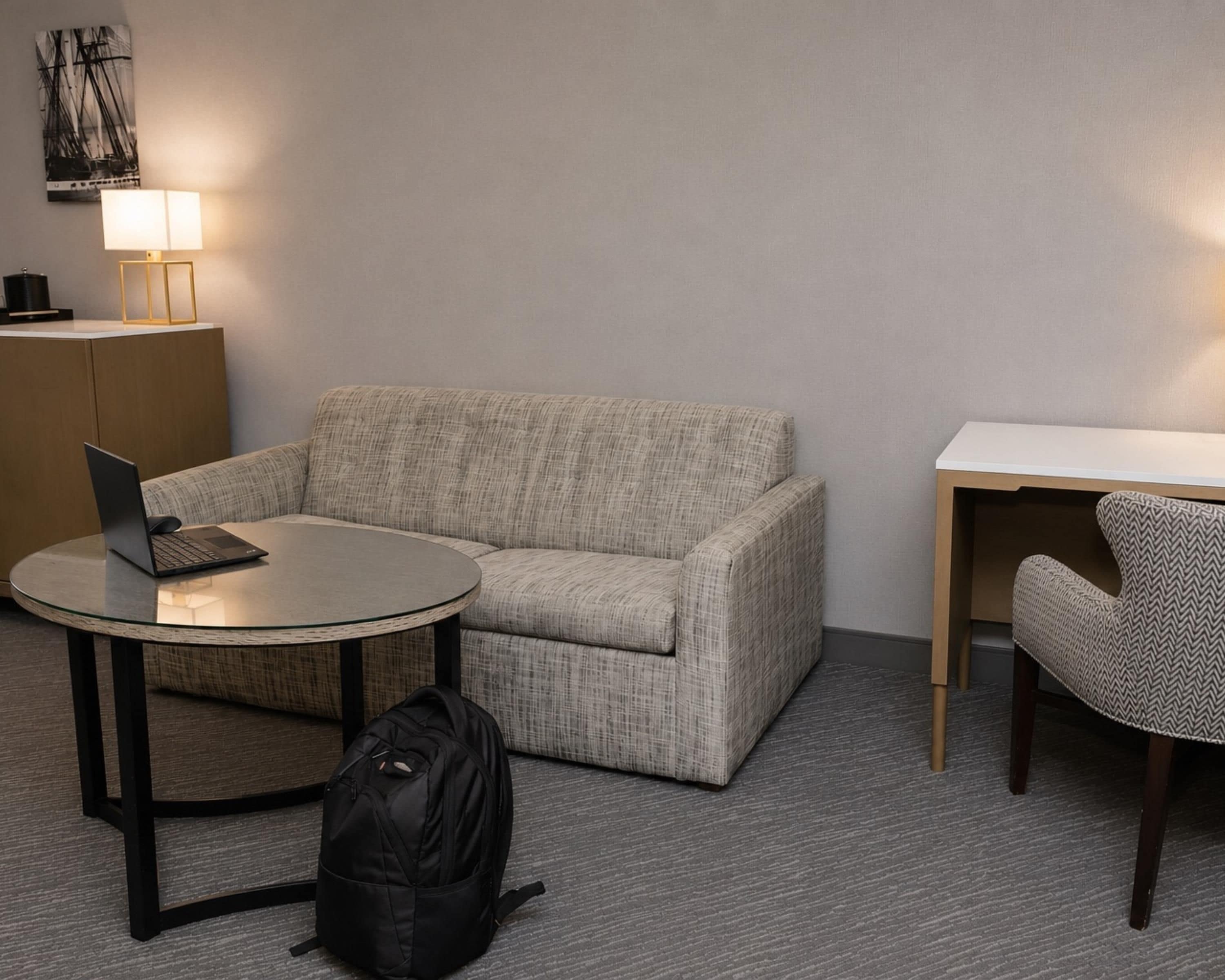 A hotel room with a sofa, a small round table with a laptop, a chair, a desk, and a lamp.