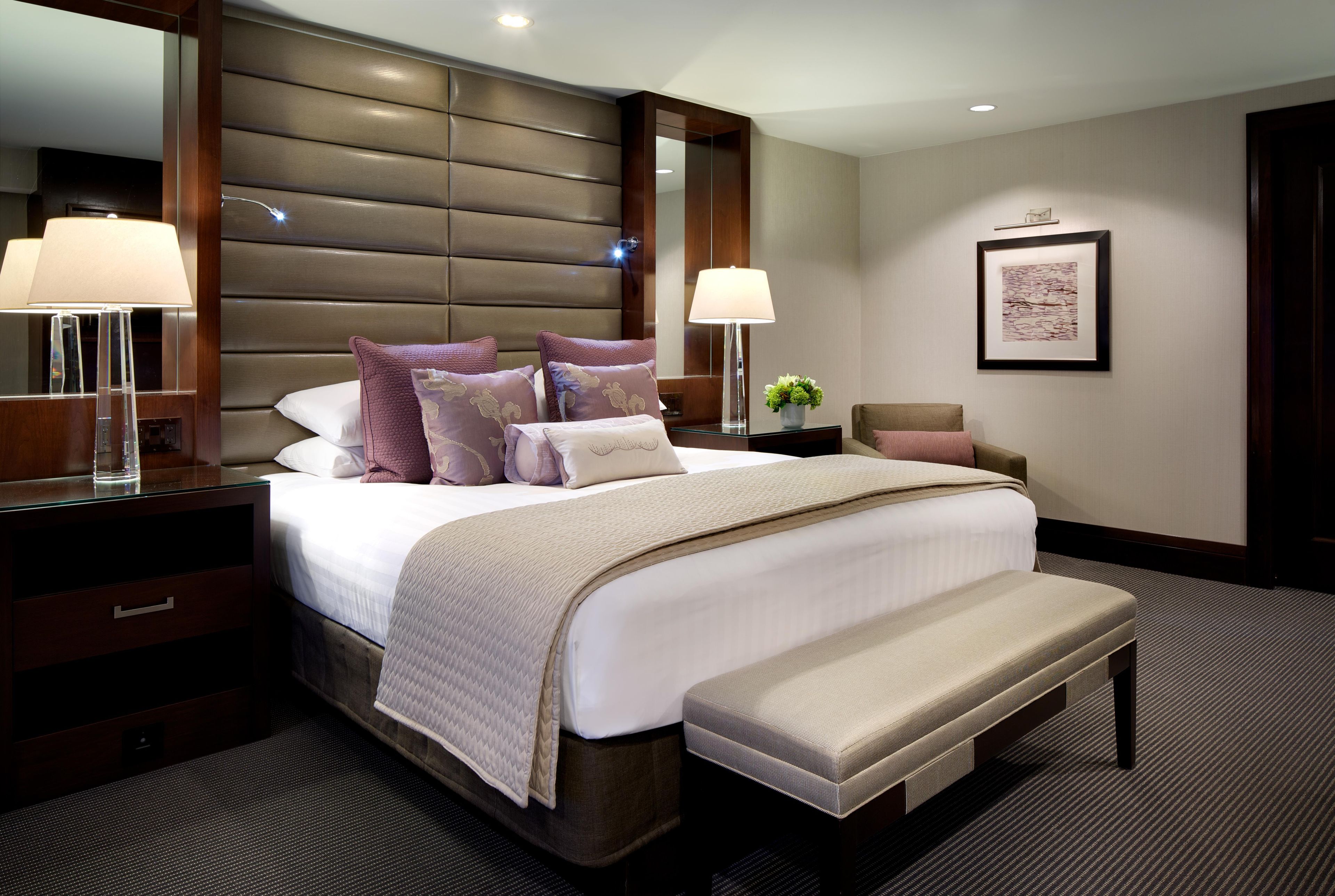 A hotel room with a large bed, bedside lamps, a bench, a nightstand, a mirror, and a framed picture on the wall.
