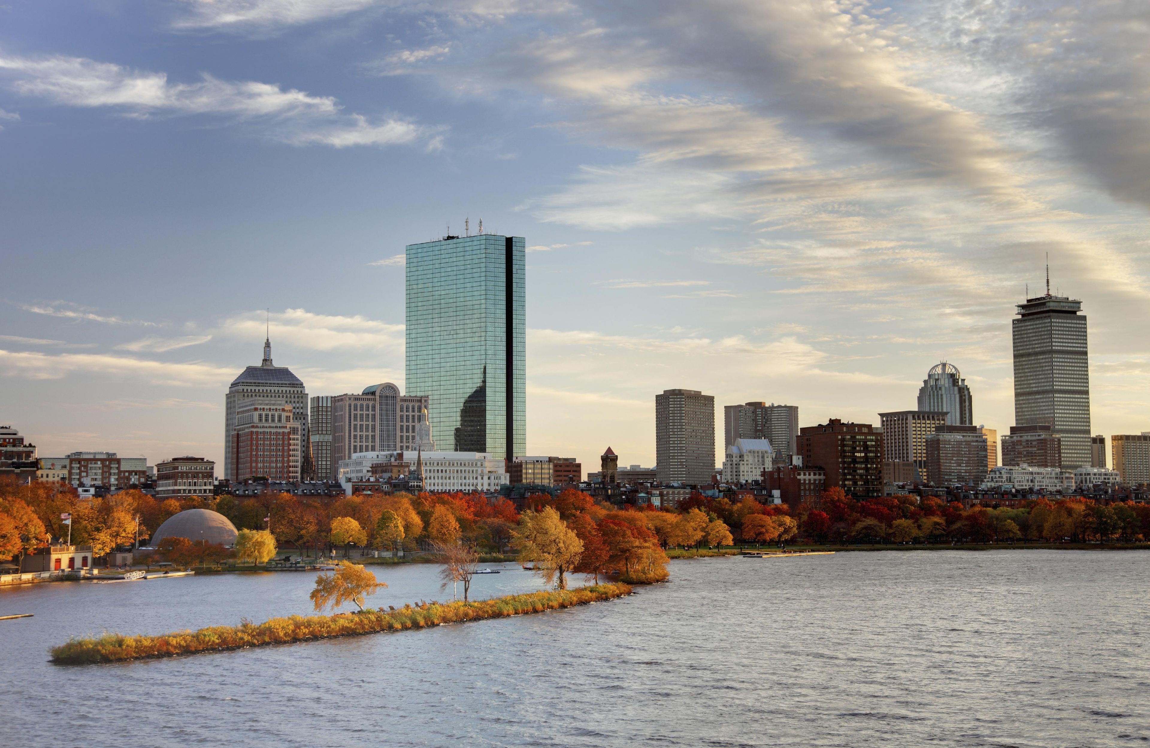 A city skyline with various buildings, including a prominent skyscraper, across a body of water with autumn foliage.