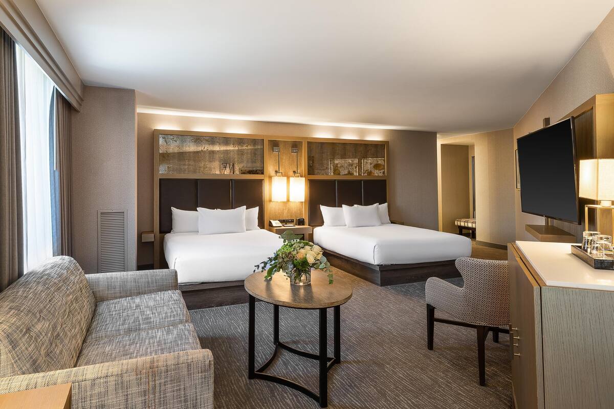 A hotel room with two beds, a sofa, a small table with a vase of flowers, a TV, and a desk with a lamp.