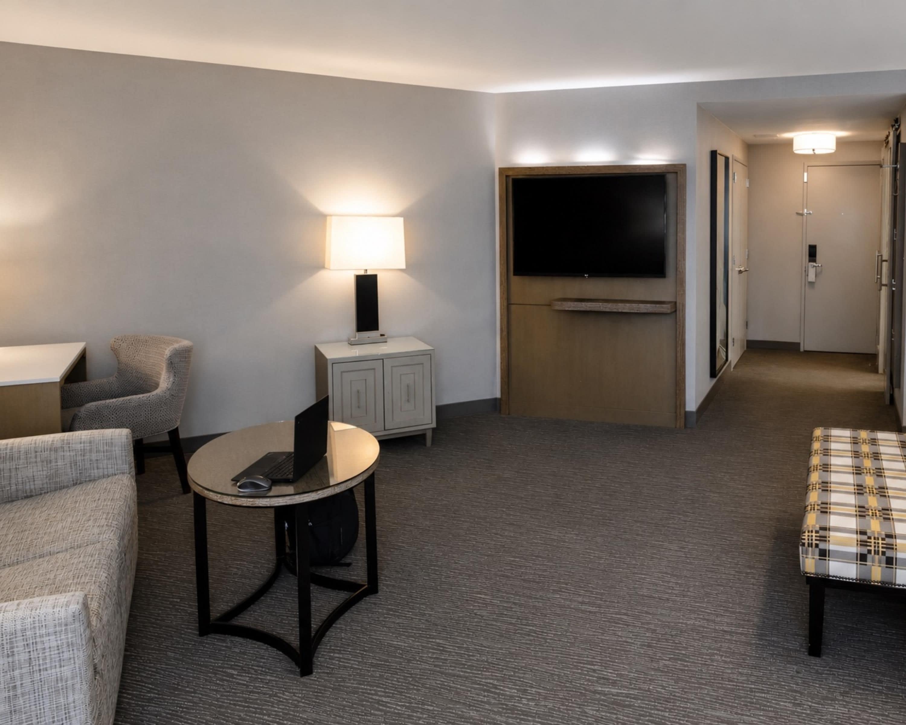 A hotel room with a flat-screen TV mounted on a wall, a small table with a laptop, a chair, a sofa, and a bed.