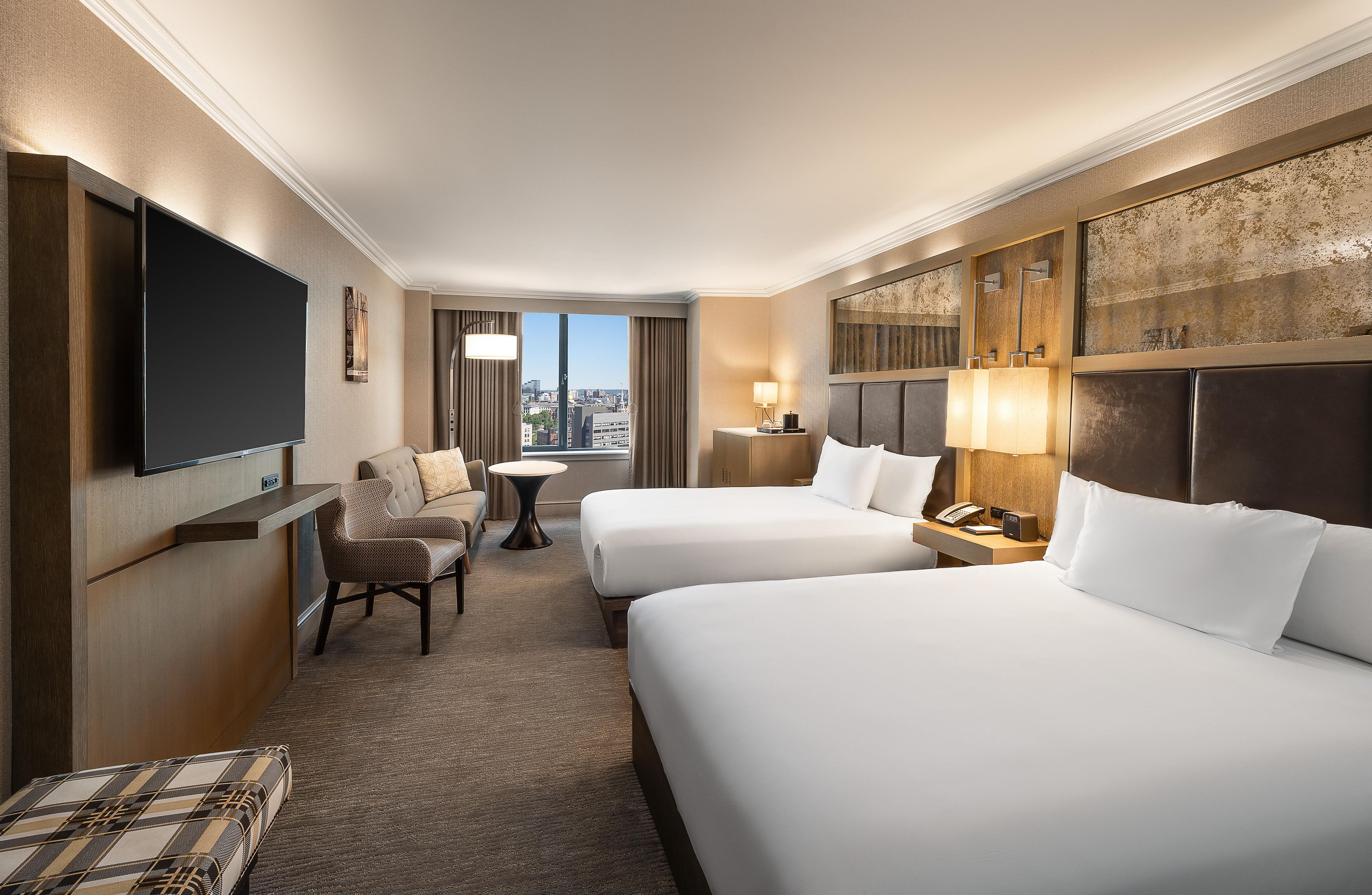 A hotel room with two beds, a TV, a desk, a chair, and a view of the city.