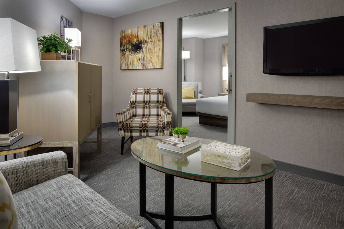 A hotel room with a glass table, a plaid armchair, a sofa, a television, and a painting on the wall.