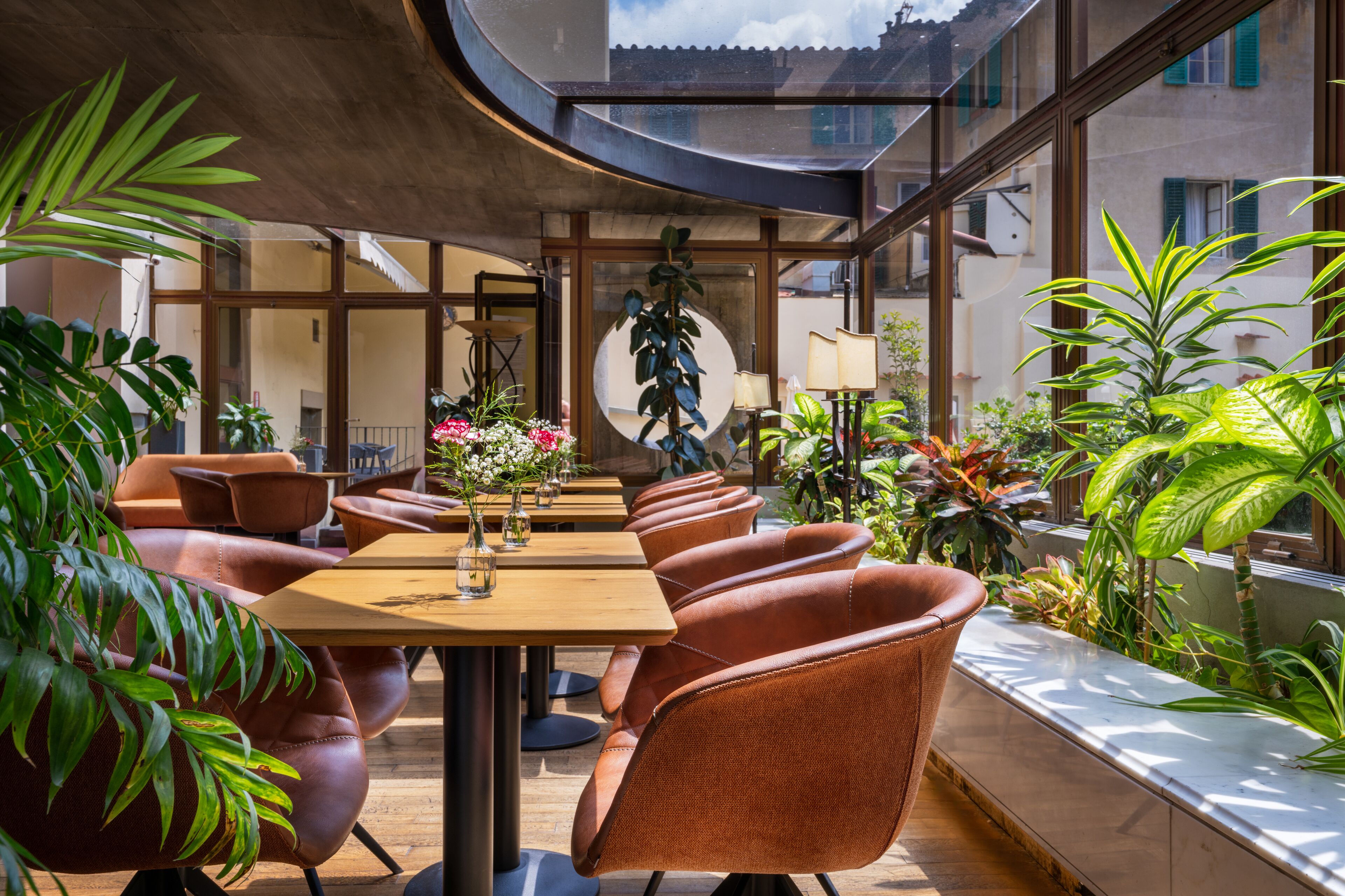 Una sala da pranzo moderna e luminosa, con sedie in pelle e tavoli, circondata da piante in vaso e ampie finestre.