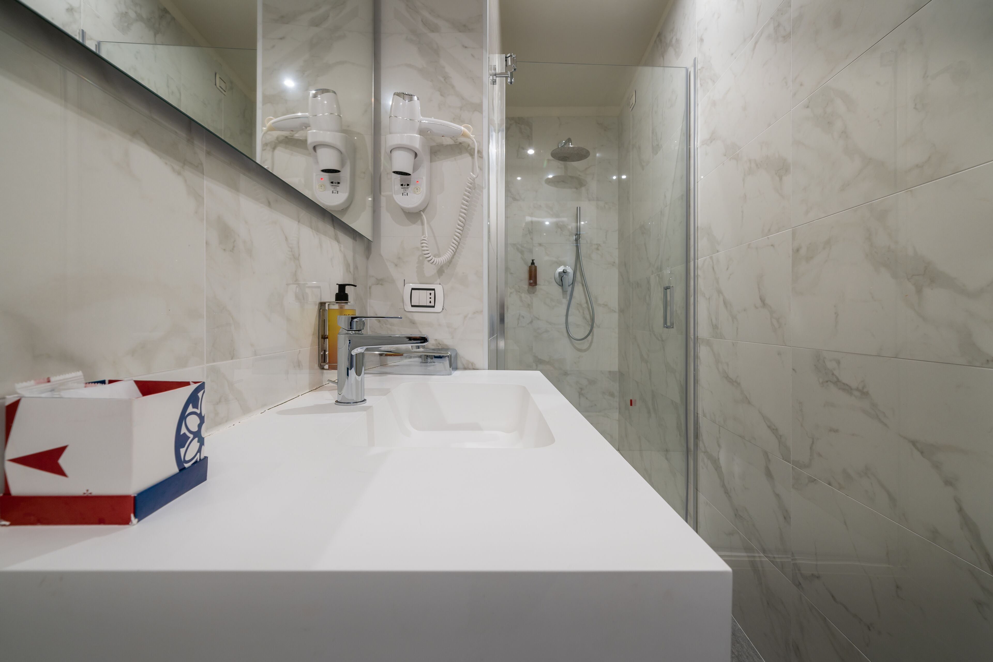 A modern bathroom with a white sink, a handheld showerhead, and a hairdryer mounted on the wall.