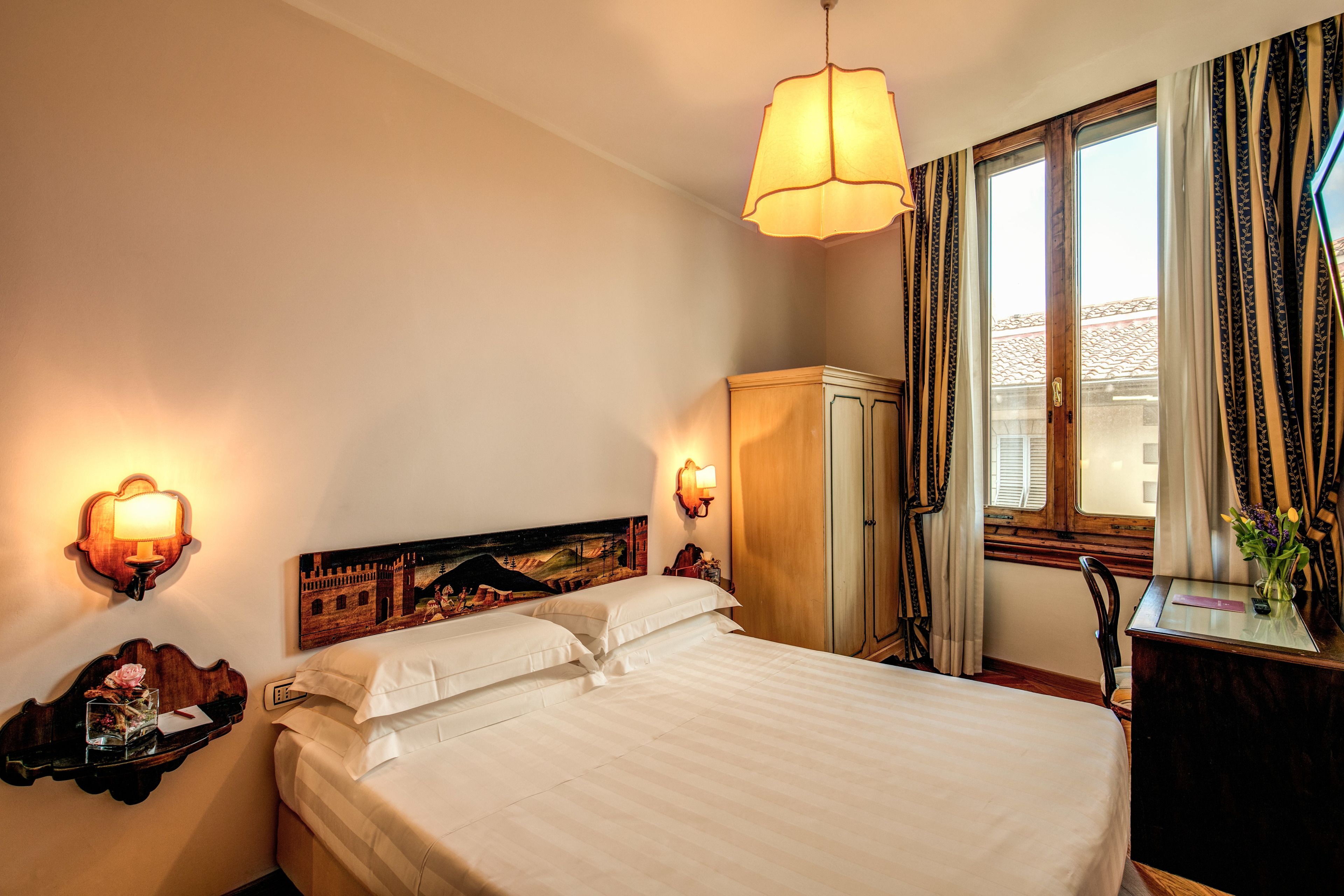 Een hotelkamer met een bed, bedlampjes, een bureau met stoel, een kledingkast en een raam met gordijnen.