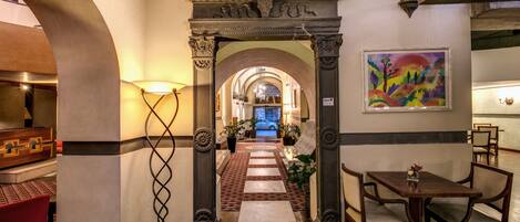 Un vestíbulo de hotel con un arco central, un comedor con mesas y sillas de madera, y un cuadro llamativo en la pared.