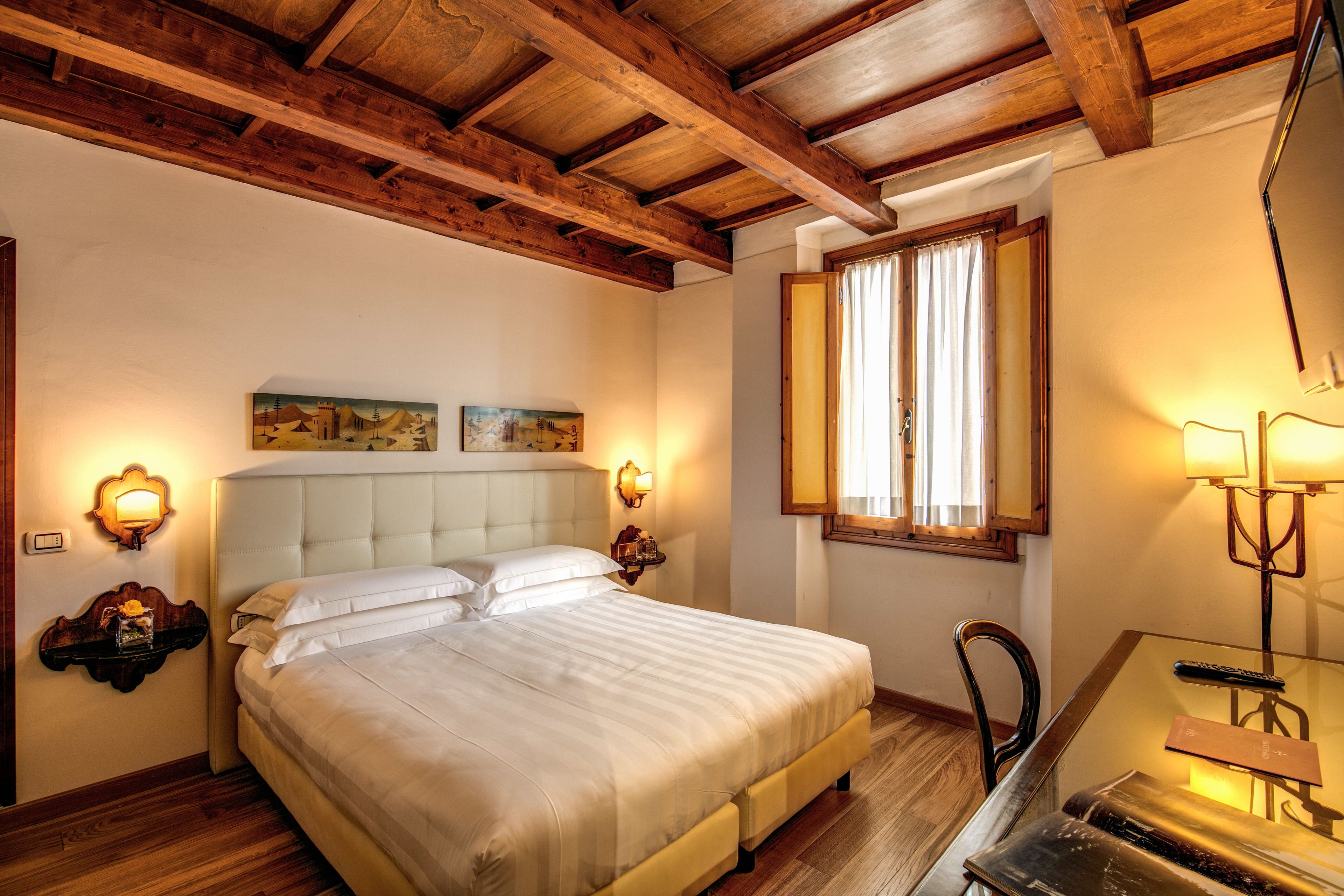 A bedroom with a large bed, wooden ceiling beams, a desk with a chair, and a window with curtains.