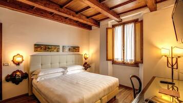 A bedroom with a large bed, wooden ceiling beams, a desk with a chair, and a window with curtains.