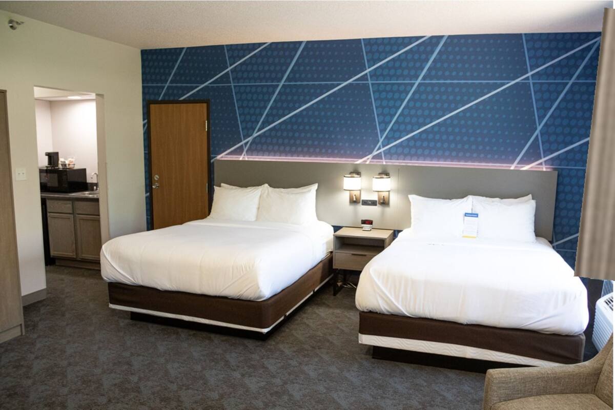 A hotel room with two beds, a desk, a chair, and a kitchenette.