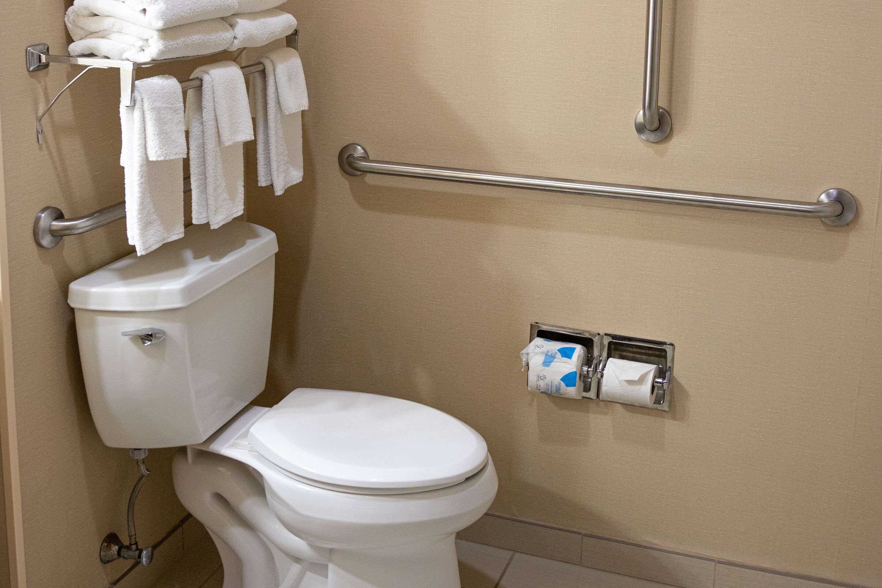 A bathroom with a white toilet, a towel rack with folded towels, a grab bar, and a paper towel dispenser.