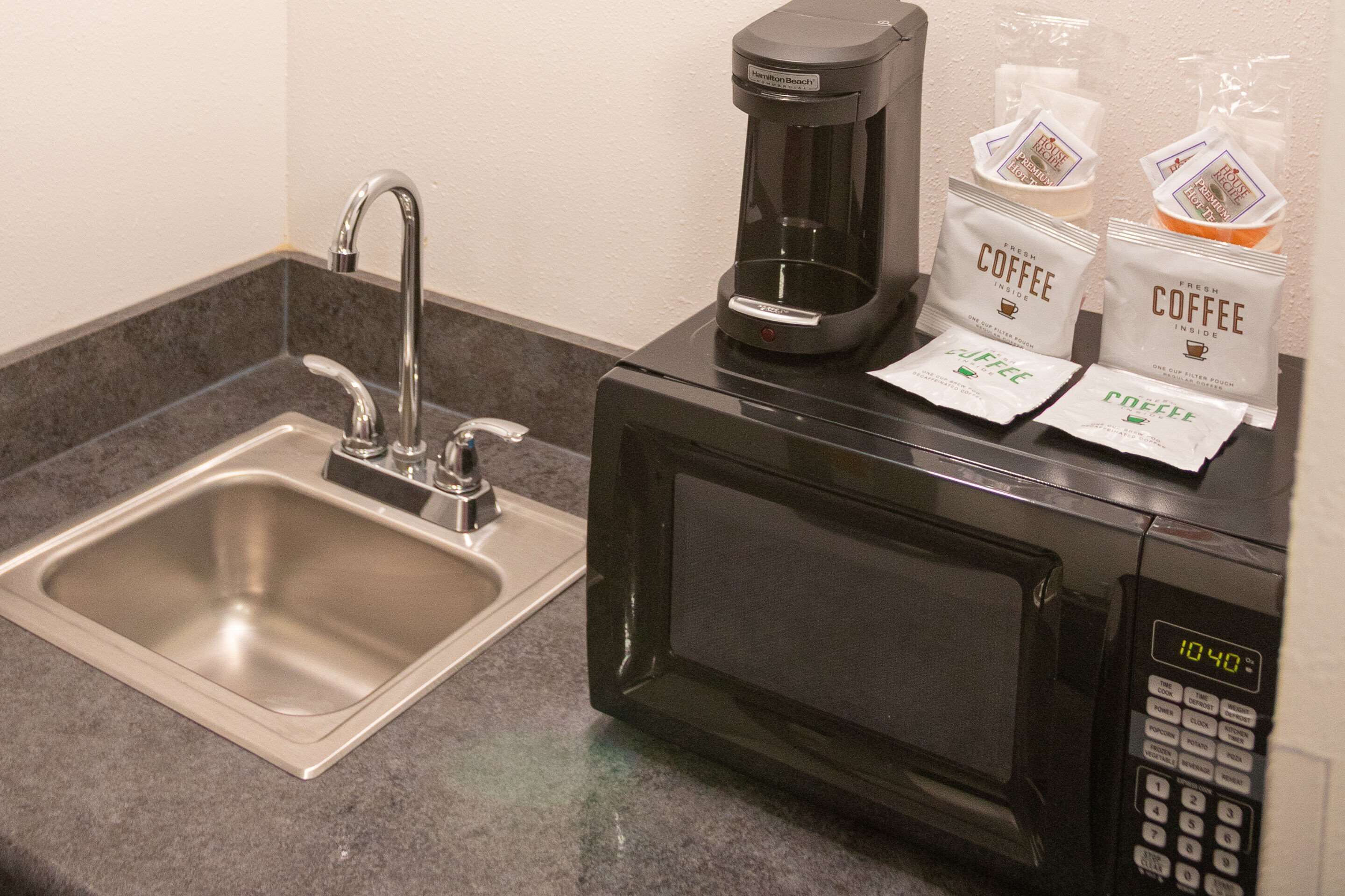 Una kitchenette con un lavamanos de acero inoxidable, una cafetera negra y dos cápsulas de café sobre el microondas.