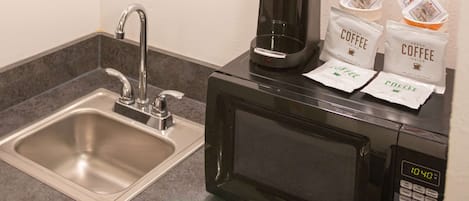 Una kitchenette con un lavamanos de acero inoxidable, una cafetera negra y dos cápsulas de café sobre el microondas.