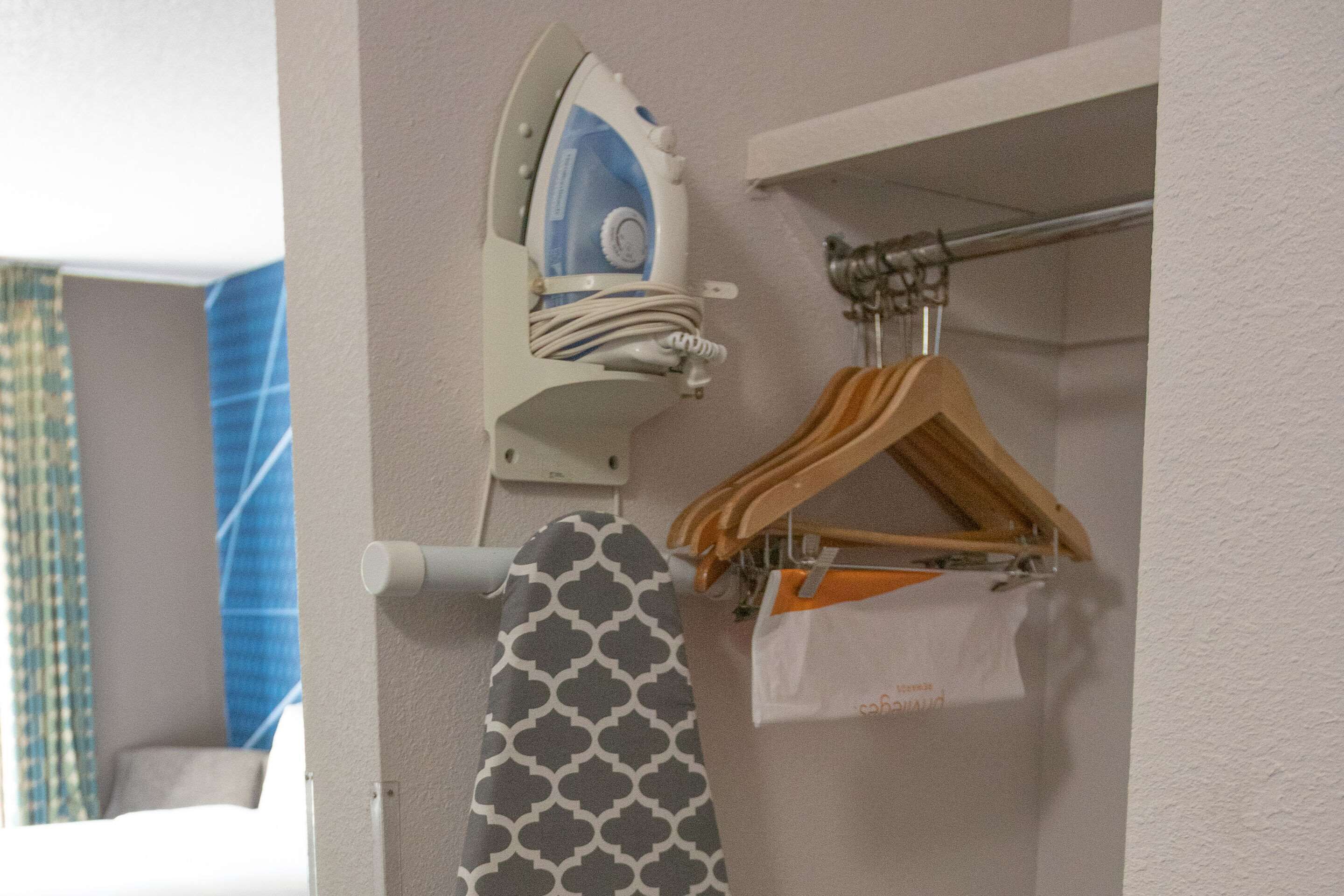 A wall-mounted ironing board with a patterned ironing board cover, a hanging wooden hanger, and a white plastic bag.