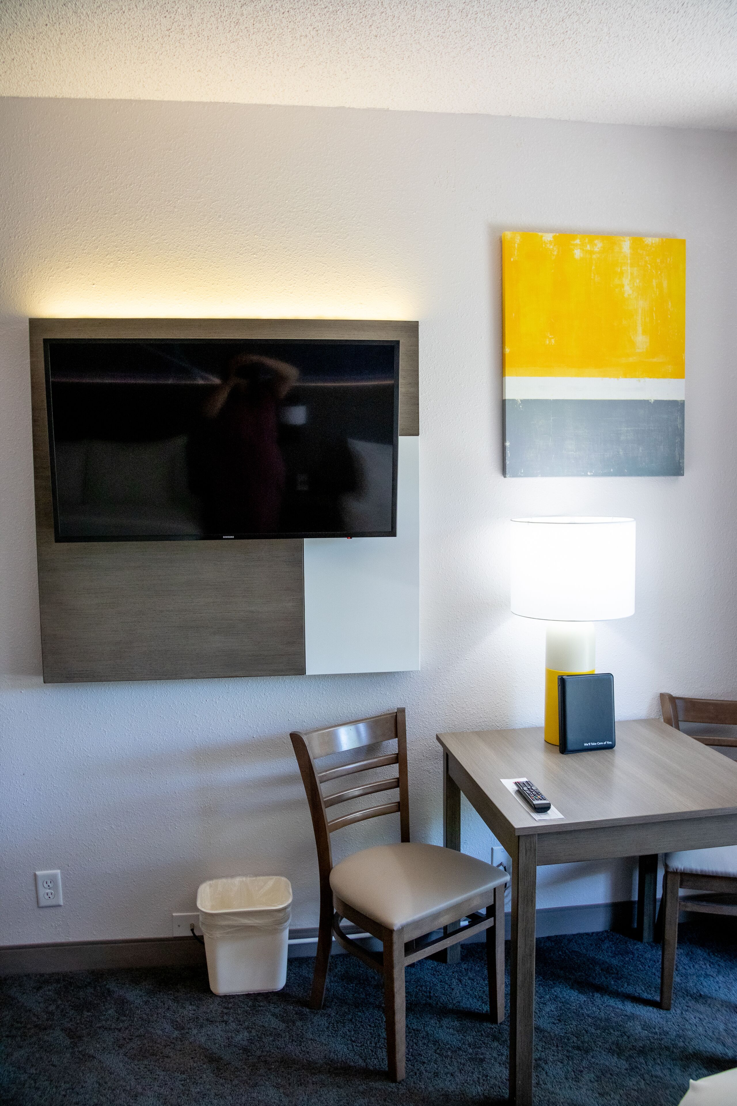 Habitación de hotel con una televisión de pantalla plana montada en la pared, una mesa con sillas, una lámpara y un cesto de basura.